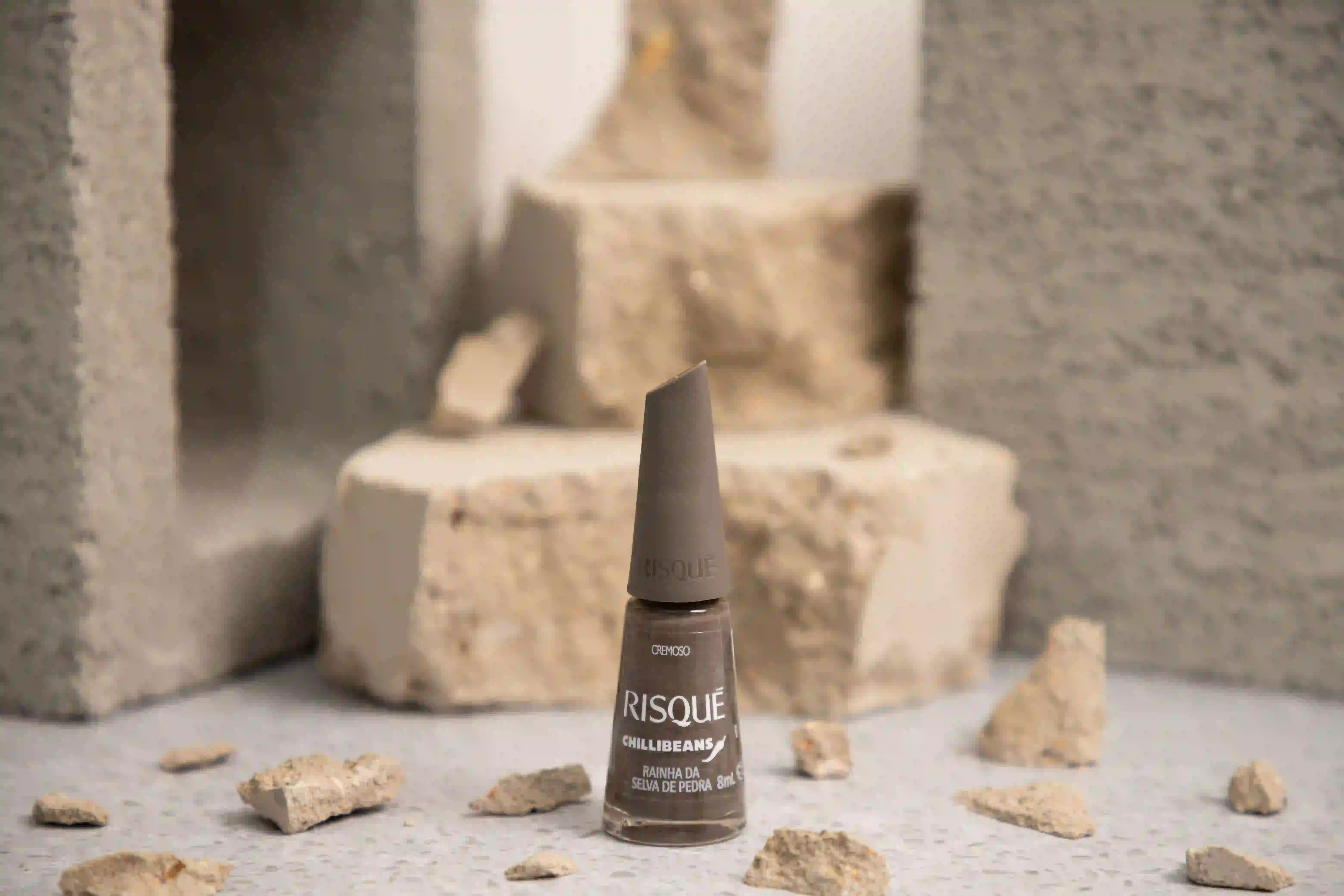 Esmalte Risqué 'Rainha da Selva de Pedra' (marrom acinzentado) sobre blocos e pedras beges, fundo cinza.