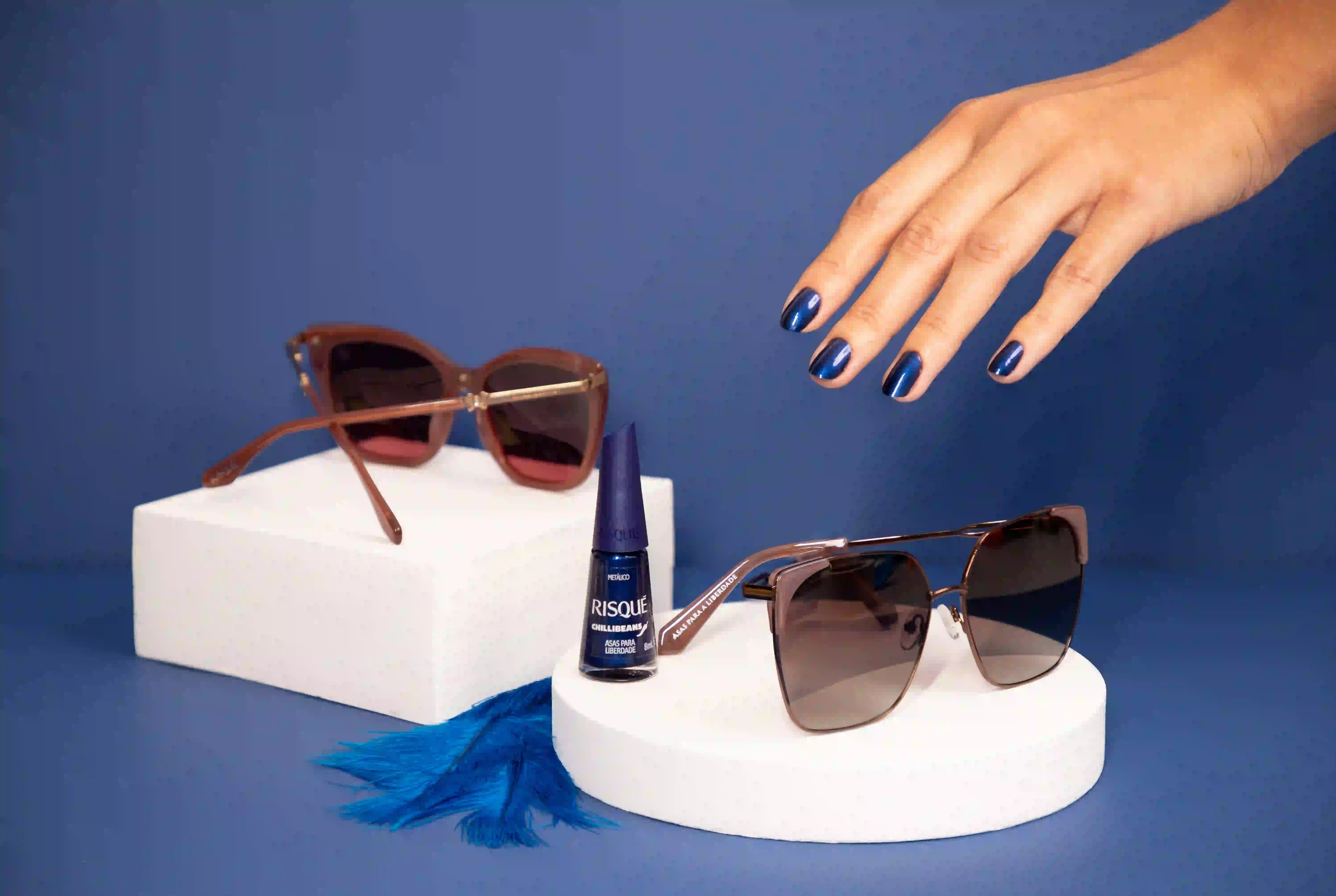Mão com unhas pintadas de azul metálico Risqué. Esmalte 'Asas para Liberdade' entre dois óculos de sol e penas azuis