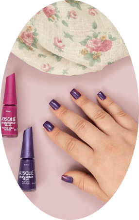 Mão com unhas roxas. Ao lado, esmalte Risqué roxo (Lágrimas de Vênus) e top coat pink, com tecido floral. Imagem oval.