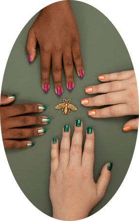 Quatro mãos com unhas pintadas de esmaltes rosa, laranja, verde metálico/glitter em volta de broche de abelha dourada.